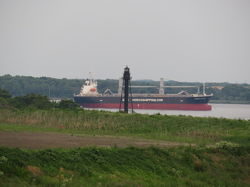 Small black skeletal lighthouse tower standing on a grassy riverbank with a large cargo ship passing on the Delaware River behind it, and a forested shore in the background. Photo by darlingtrk (https://commons.wikimedia.org/wiki/File:Bellevue_Rear_Range_Lighthouse_(28809856398).jpg ), CC BY 2.0 (https://creativecommons.org/licenses/by/2.0 ), via Wikimedia Commons.