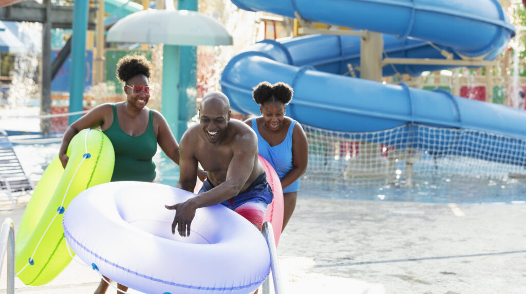 An African-American family with a 12 year old girl having fun at a water park. They are carrying inflatable rings, ready to float on the lazy river. Giant water slides are in the background