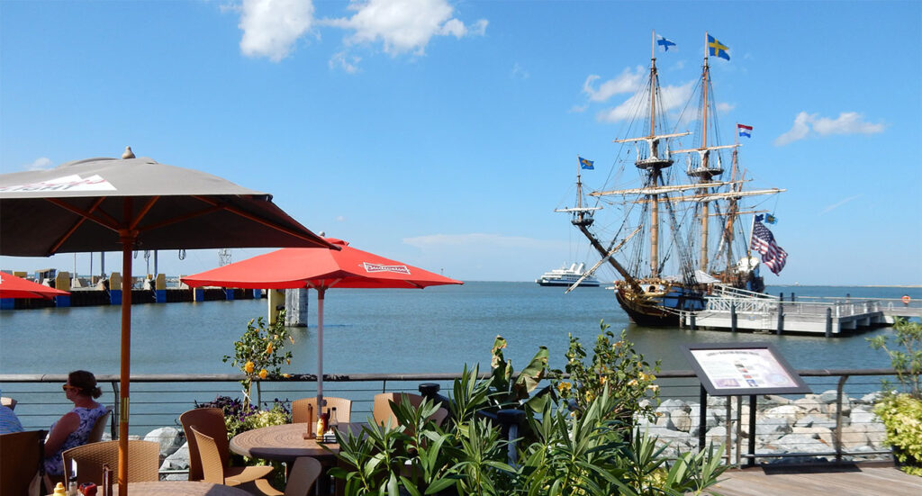 Lewes, DE: A waterfront restaurant patio with red umbrellas and a wooden deck. In the harbor, a large, historic wooden tall ship, the Kalmar Nyckel, is docked under a blue sky with white clouds.