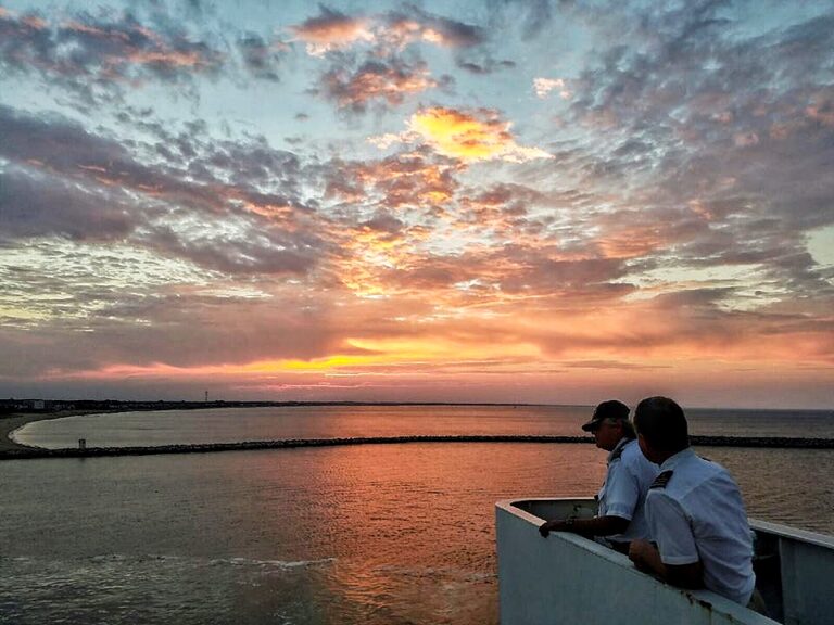 Two Cape May-Lewes Ferry Captains in uniform observing a vivid sunset over the Delaware Bay from the upper deck, highlighting the scenic journey.