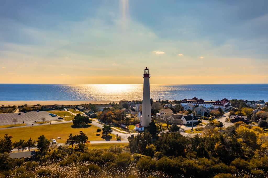 Aerial view of Cape May, NJ showing the lighthouse, surrounding town, sandy beach, and Delaware Bay at golden hour.
