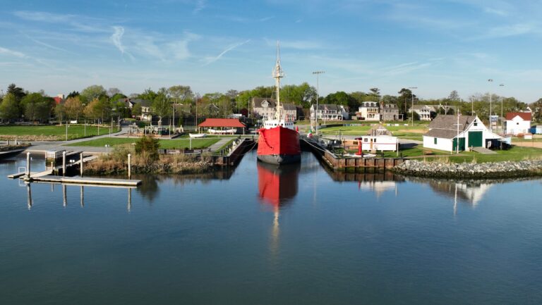 A red lightship permanently docked along the Lewes canal, reflected in calm water, with historic buildings and green lawns visible along the shoreline.