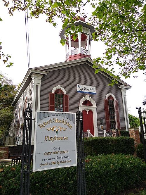 Exterior of the Robert Shackleton Playhouse, home of Cape May Stage, featuring a Victorian-era gray building with red trim, arched windows, and a decorative cupola. Photo by WhisperToMe (https://commons.wikimedia.org/wiki/File:Cape_May_Theatre0.jpg) , CC BY-SA 4.0 (https://creativecommons.org/licenses/by-sa/4.0) , via Wikimedia Commons
