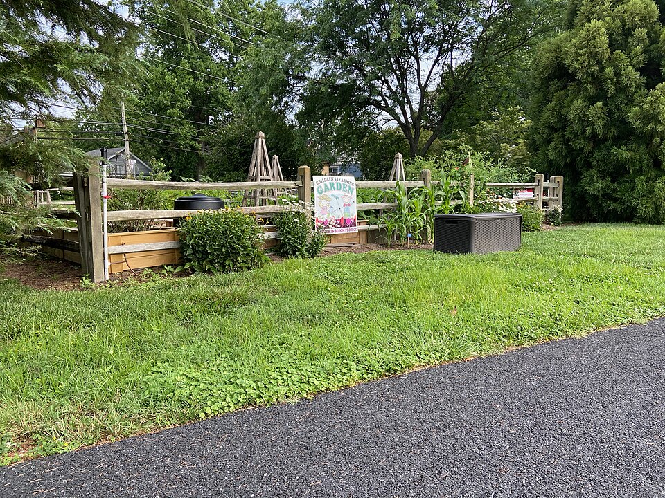 The paved multi-use Georgetown-Lewes trail running alongside a fenced Children's Learning Garden with raised beds and trellises in Lewes, DE. Photo by Nolabob (https://commons.wikimedia.org/wiki/File:Children%27s_Learning_Garden.jpg), CC BY-SA 4.0 (https://creativecommons.org/licenses/by-sa/4.0) , via Wikimedia Commons.