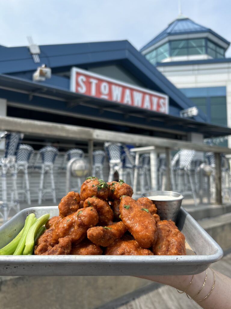 A hand holds a metal tray of sauced chicken wings with celery and dipping sauce outside Stowaways restaurant in Cape May, NJ, with the Stowaways sign and outdoor bar seating visible in the background