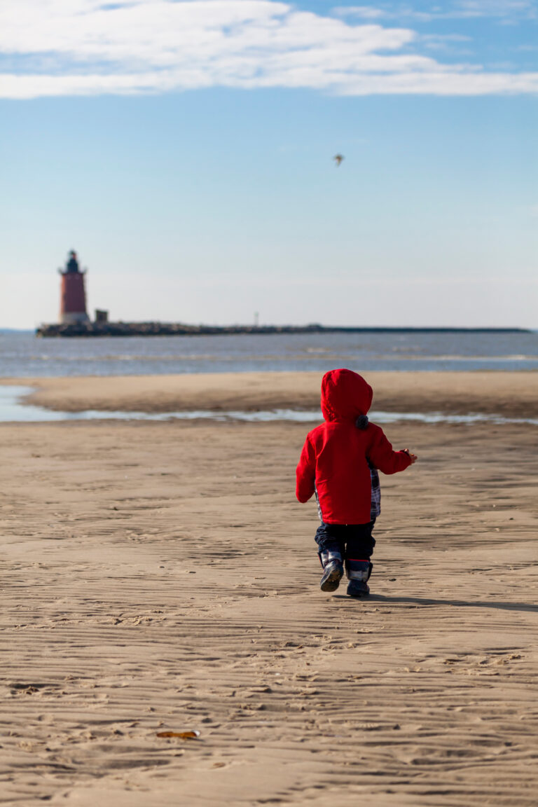 A toddler in a red jacket runs along a sandy beach with a red and black lighthouse visible in the background.