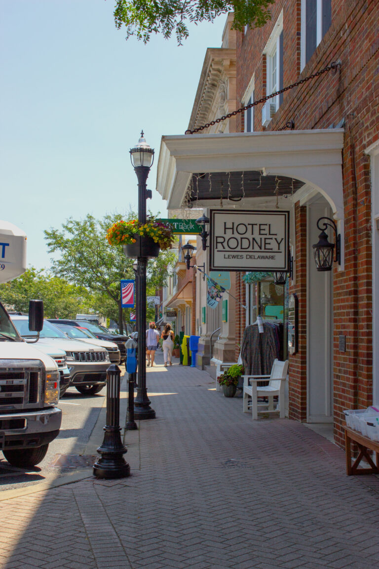 A brick sidewalk lined with lamp posts and storefronts in historic downtown Lewes, Delaware, with the Hotel Rodney sign visible on a brick building.