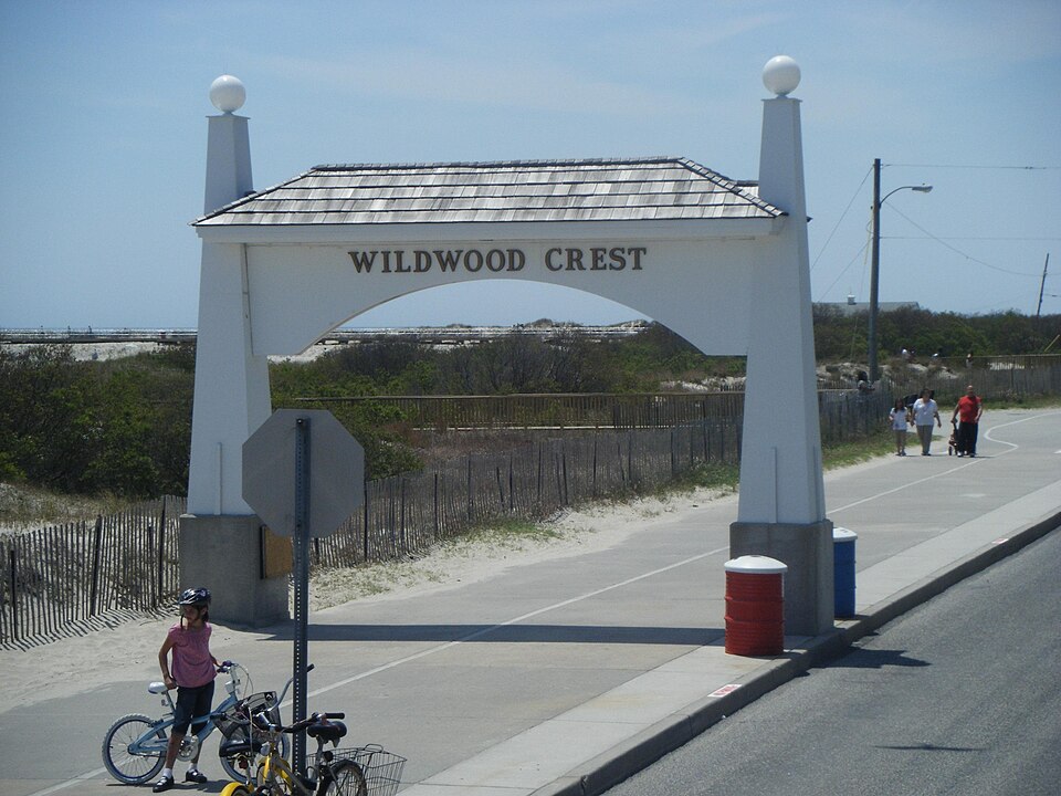 Wildwood Crest entrance arch with cyclists and beachgoers on the paved path, beach dunes and coastal access visible in the background. Photo by Dough4872 (https://commons.wikimedia.org/wiki/File:Wildwood_Crest_arch.JPG) , Public domain, via Wikimedia Commons.
