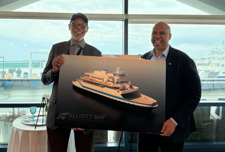 Two men stand in front of a large expanse of windows with a Ferry visible in the background while the men, Joel Coppadge on the left, and Senator Cory Booker on the right, hold a graphic representation of the hybrid Ferry currently being built for the Cape May - Lewes Ferry on August 28, 2026