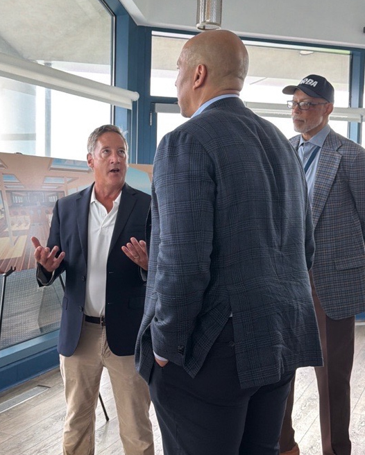 Heath Gehrke, Director of Ferry Operations talks to Senator Cory Booker while Joel Coppadge looks on. Heath is sharing a display of interior renderings of the new ferry being constructed in Rhode Island.