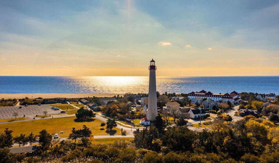 Aerial view of Cape May, NJ showing the lighthouse, surrounding town, sandy beach, and Delaware Bay at golden hour.