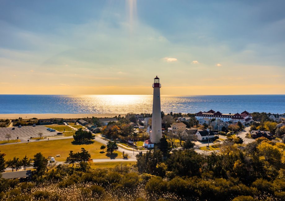 Aerial view of Cape May, NJ showing the lighthouse, surrounding town, sandy beach, and Delaware Bay at golden hour.